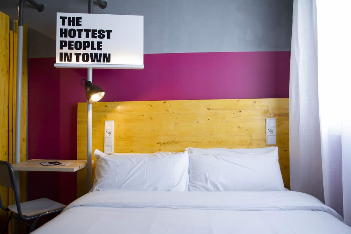 Quarto de hotel com cabeceira de madeira, cama com roupa de cama branca, mesa de cabeceira com abajur e um letreiro acima da cama que diz 'THE HOTTEST PEOPLE IN TOWN'.