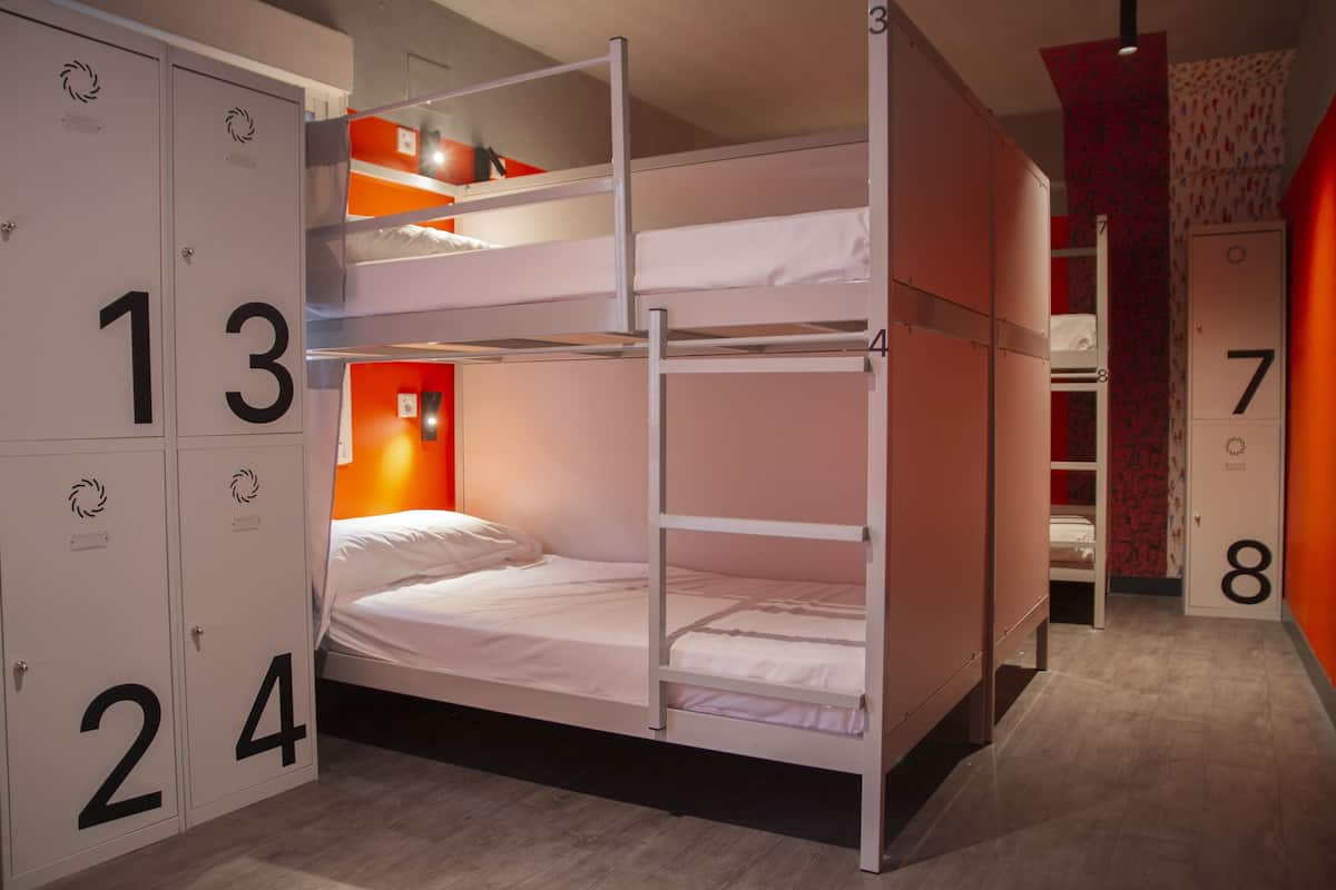 Quarto com beliche, armários individuais numerados de 1 a 8, uma parede com destaque em vermelho e uma porta que dá para outro quarto.