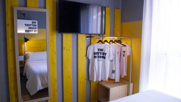 A hotel room with a bed, a mirror, a TV mounted on the wall, and a clothing rack with t-shirts.
