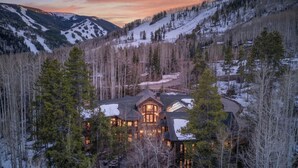 A large, multi-story lodge with a prominent central tower, surrounded by dense forest and snow-covered mountains. - Luxury Ski-in/out Estate Hot tub + Game Room Paragon (Avon)