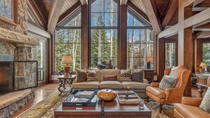 A spacious living room with a stone fireplace, large windows, and a leather armchair. - Luxury Ski-in/out Estate Hot tub + Game Room Paragon (Avon)