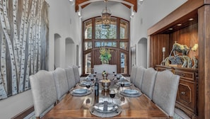 A dining room with a long table set for a meal, surrounded by high-backed chairs, with a large window and a chandelier. - Luxury Ski-in/out Estate + Game Room | Paragon (Avon)