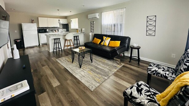 Ein modernes Wohnzimmer mit einer schwarzen Ledersofa, einem Couchtisch und einem gemusterten Sessel. Im Hintergrund ist eine Küche mit weißen Schränken und einem Kühlschrank zu sehen.