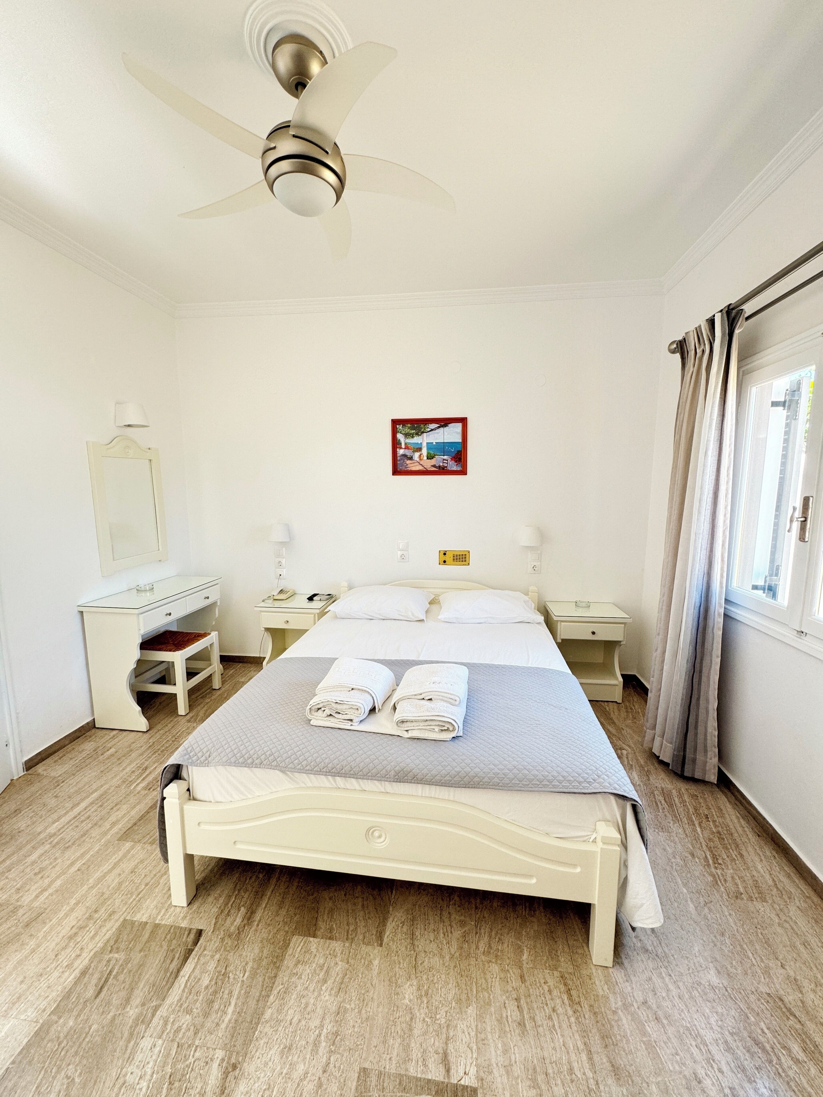 Un dormitorio ordenado con cama, mesitas de noche, escritorio, espejo y ventilador de techo.