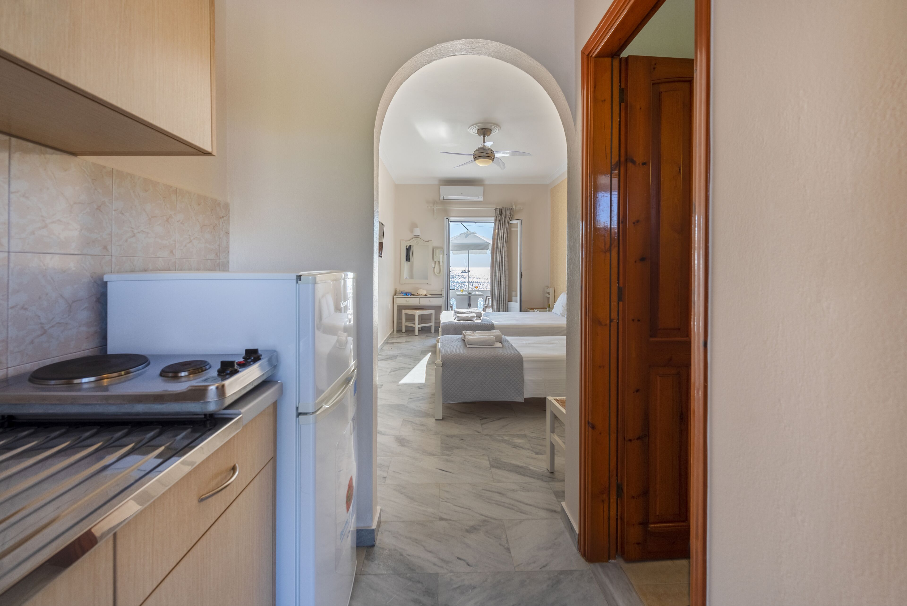 Una cocina con estufa, refrigerador y armarios de madera. Un pasillo conduce a un dormitorio con cama y ventana.