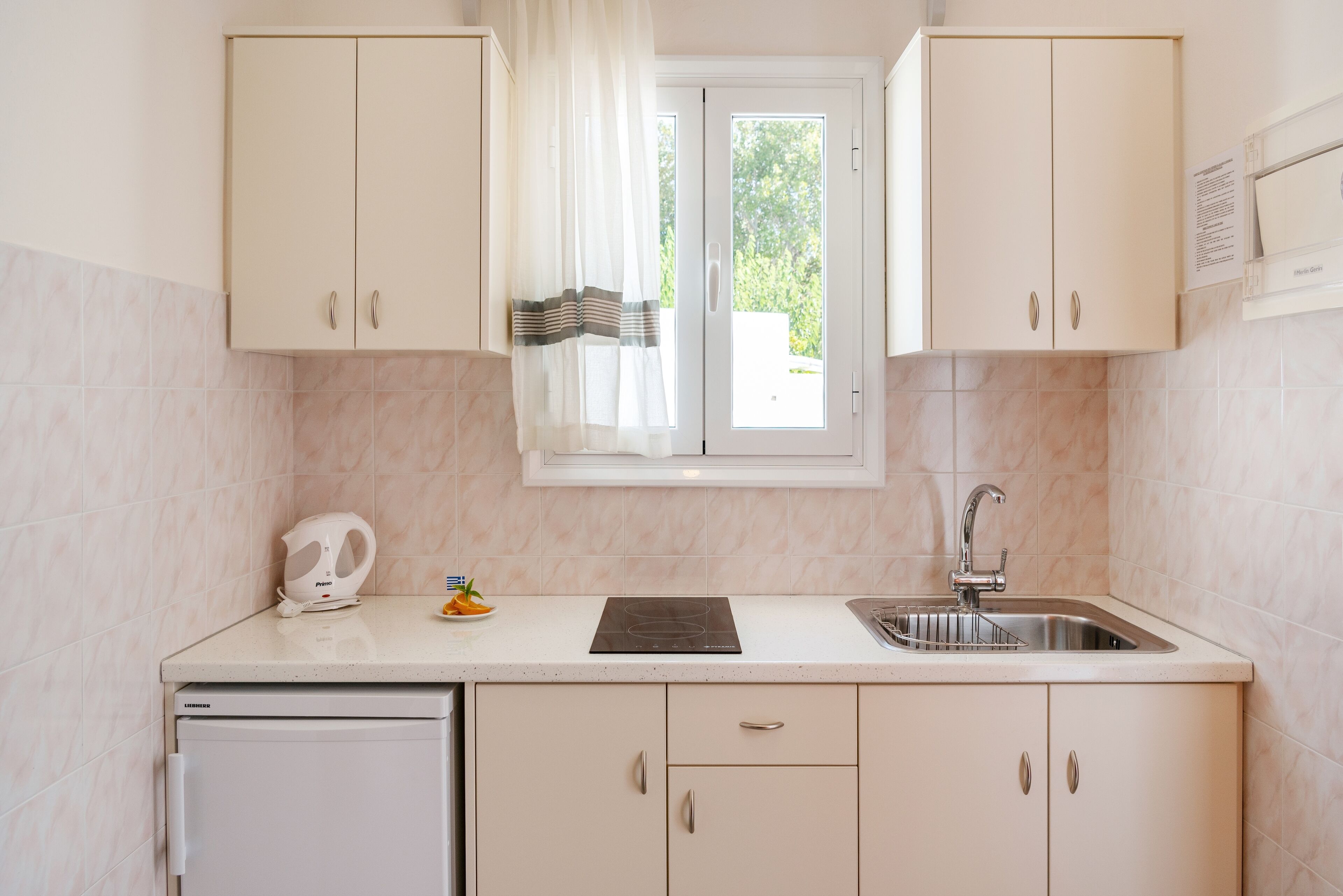 Een compacte keuken met witte kastjes, een inbouwvaatwasser, een raam met gordijn en een spoelbak.