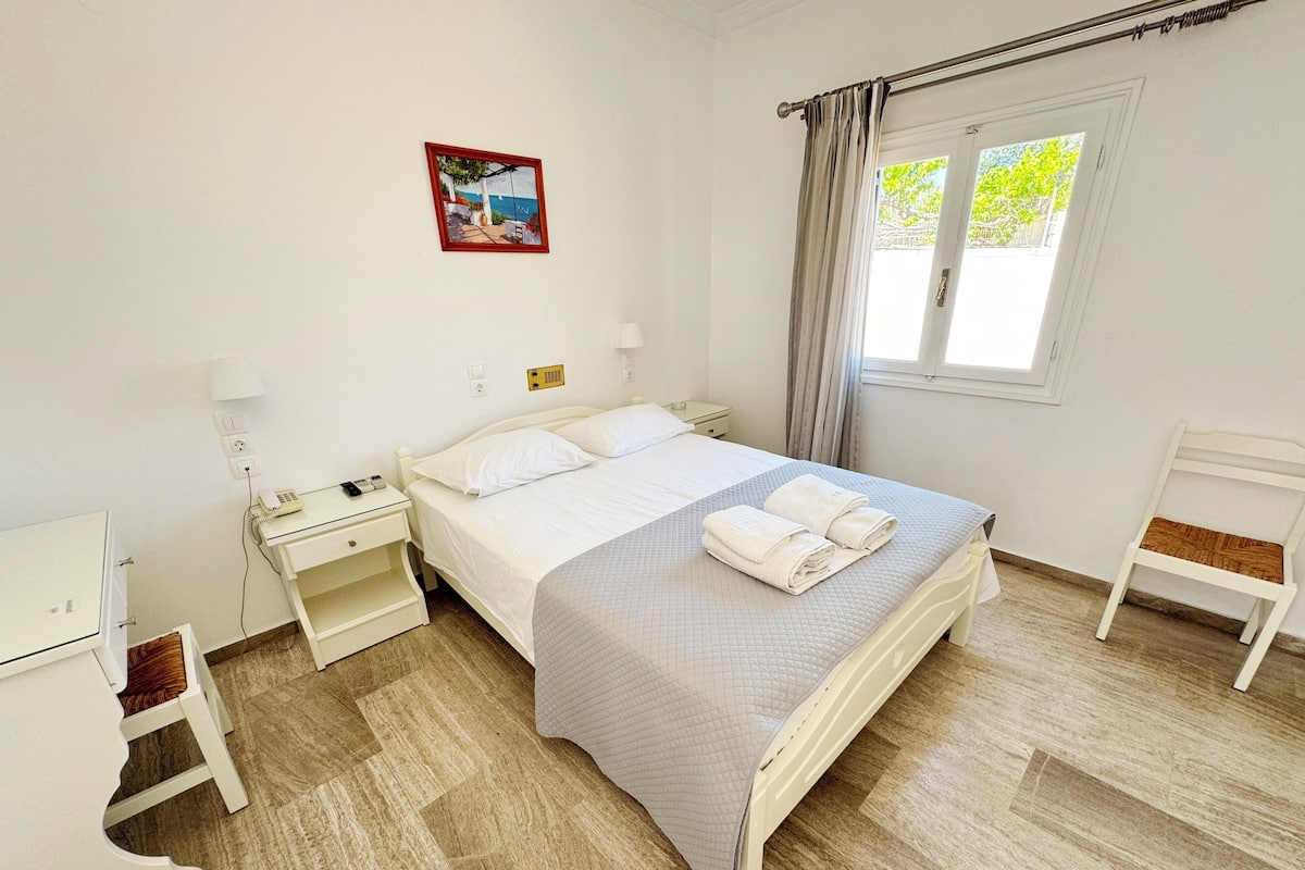Un dormitorio ordenado con cama, mesitas de noche, un escritorio y una ventana con cortinas.