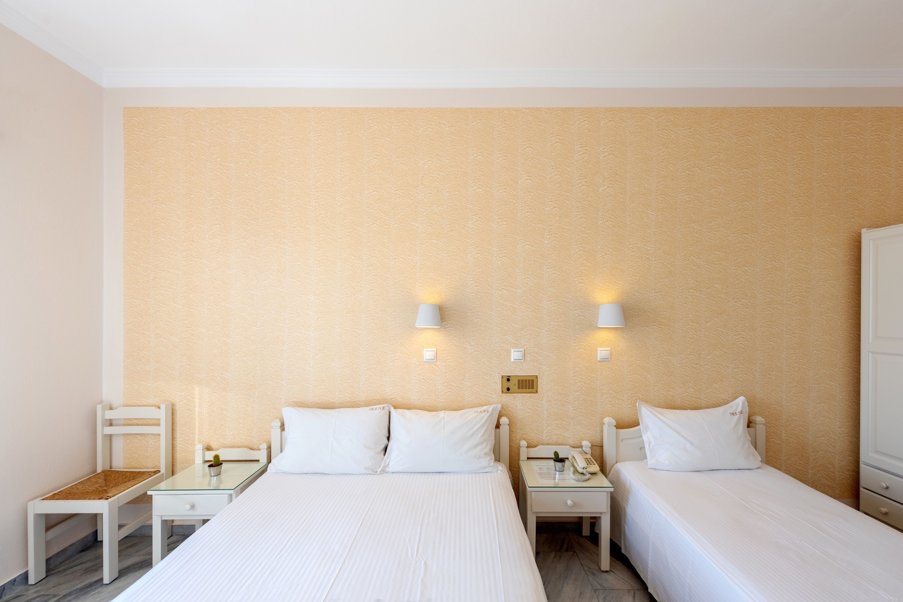 A hotel room with two single beds, a small table, a chair, and a nightstand with a lamp.