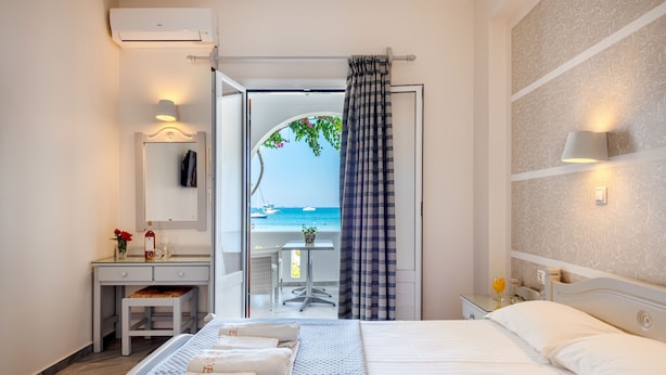 Ein Hotelzimmer mit Bett, Schreibtisch, Stuhl, kleinem Tisch mit Lampe und Meerblick durch eine offene Tür.
