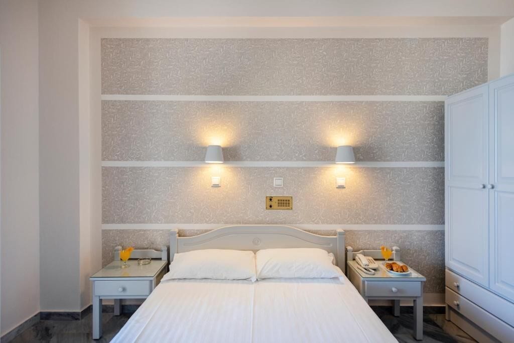 Un lit double avec du linge de lit blanc, deux tables de chevet avec des lampes, un luminaire fixé au mur et une armoire.