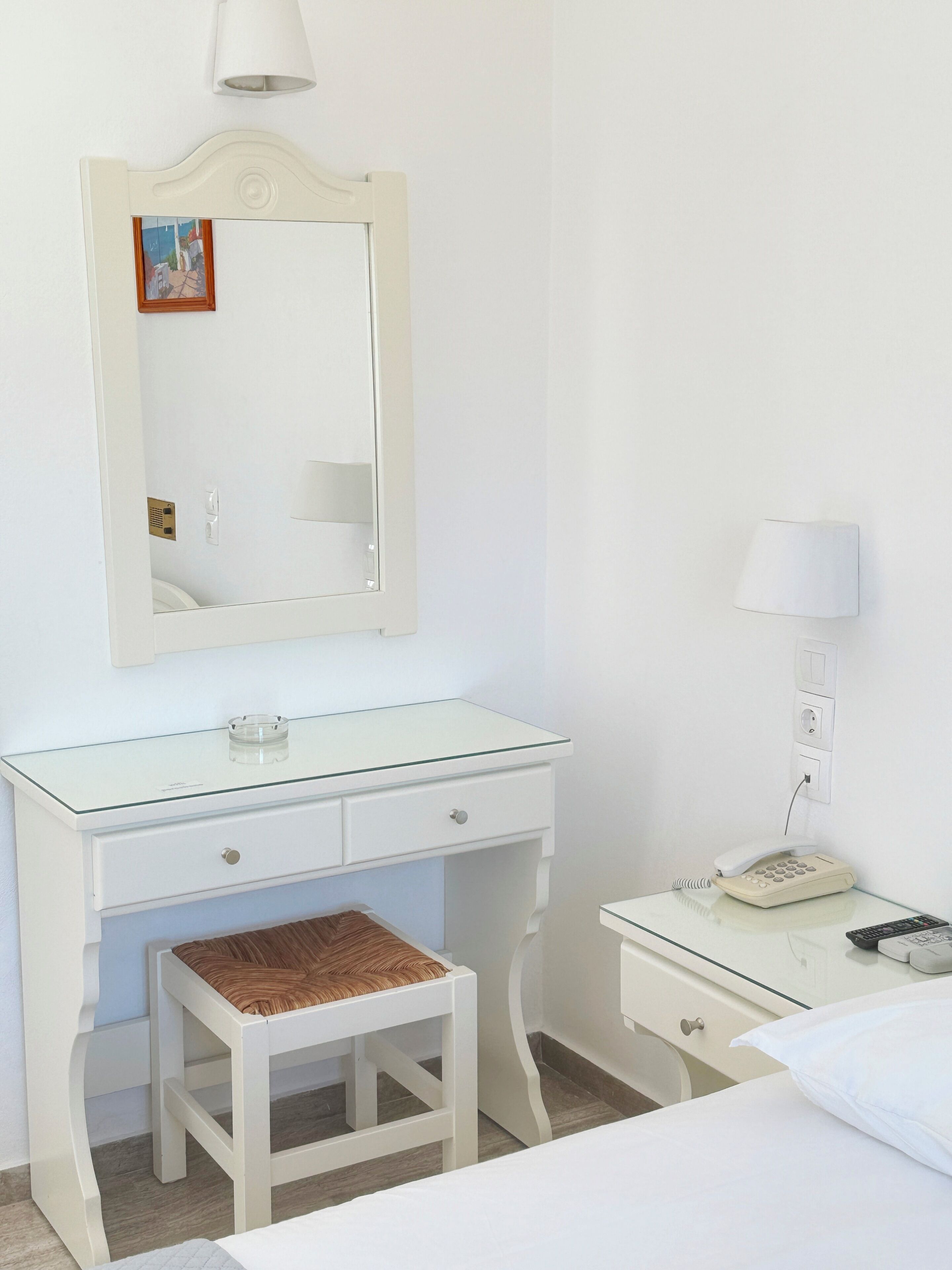 A white room with a glass-top desk, a small stool, a mirror, a lamp, a telephone, and a bed.