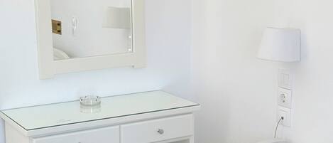 A white room with a glass-top desk, a small stool, a mirror, a lamp, a telephone, and a bed.