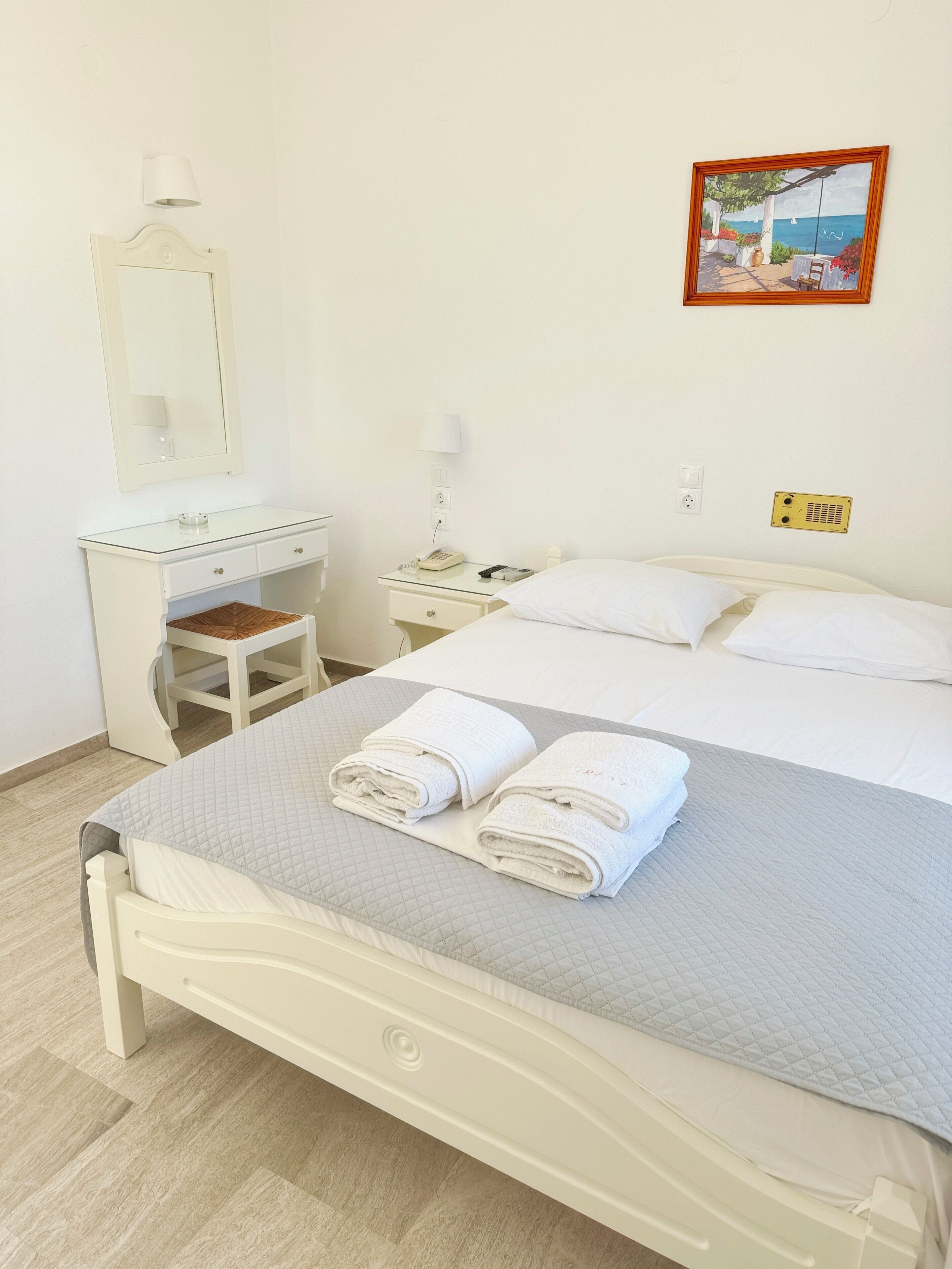 A neatly made bed with two folded towels, a bedside table with a lamp, a small desk with a chair, and a wall-mounted picture.