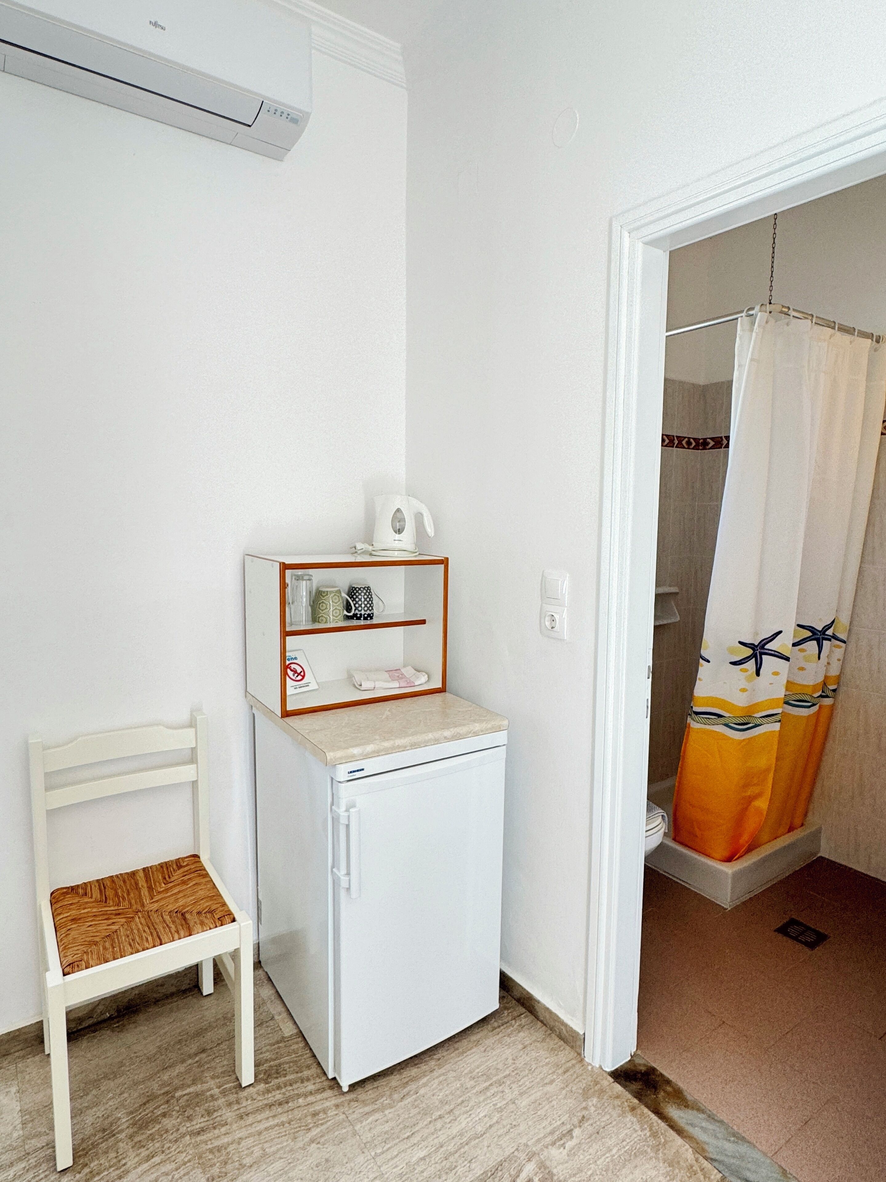Una habitación compacta con un refrigerador blanco, un pequeño estante de madera con utensilios de cocina, una silla blanca y una zona de ducha con cortina.