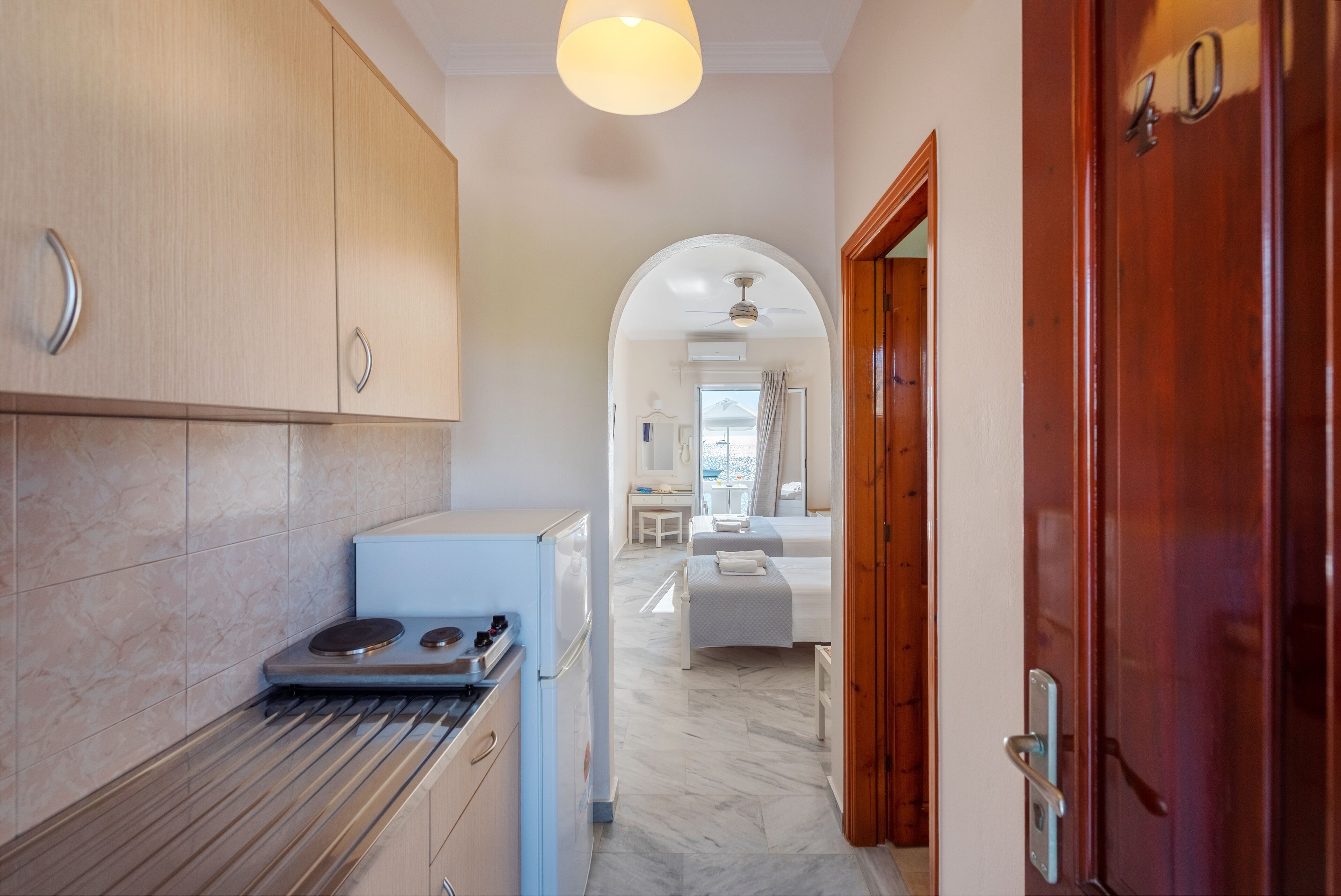 A narrow hallway with a kitchen area on the left, a wooden door on the right, and a bedroom visible through an archway in the background.