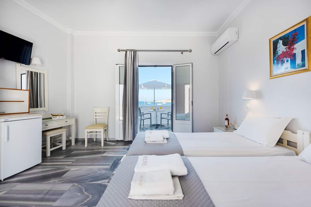 Habitación de hotel con dos camas, un escritorio, una silla y vista al mar por una puerta abierta.