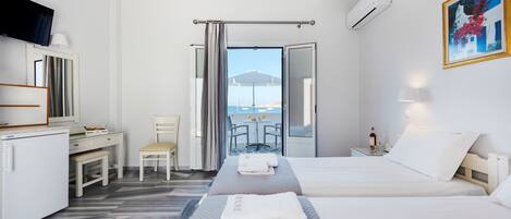 A hotel room with two beds, a desk, a chair, and a view of the sea through an open door.