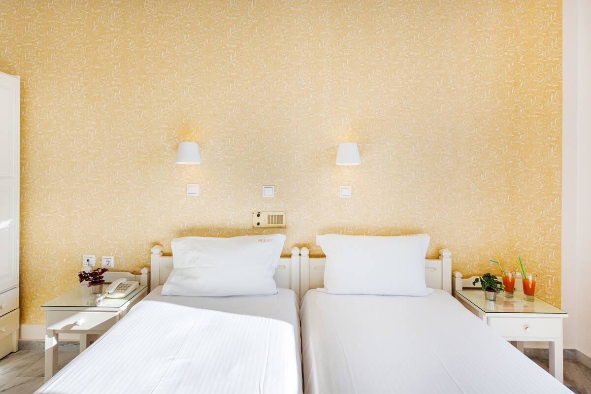 Habitación de hotel con dos camas, una mesita de noche con teléfono y planta, y una lámpara de pared.