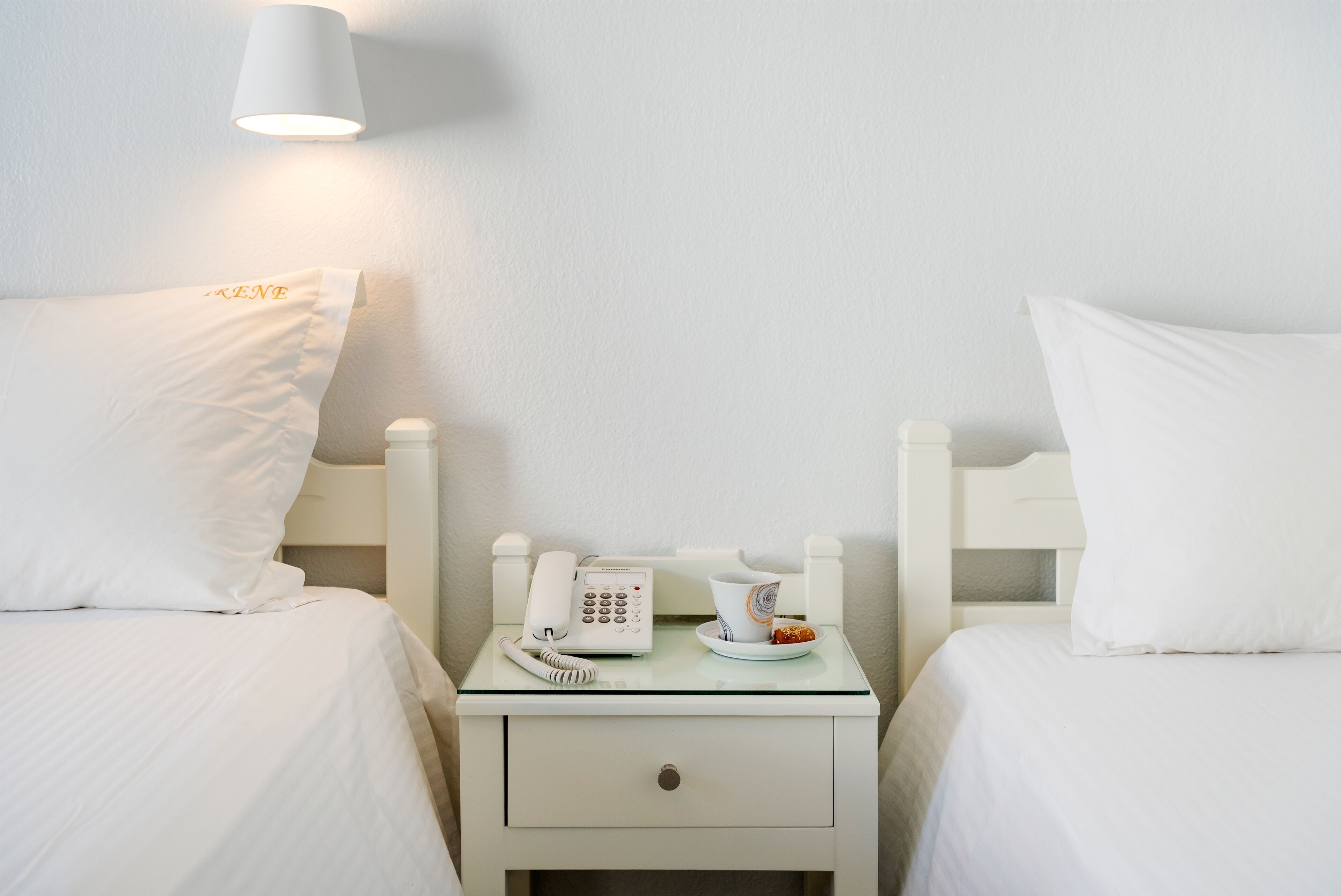 A hotel room with two beds, a bedside table with a phone, a cup, and a plate with a pastry.