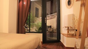 A bedroom with a bed, a sliding glass door leading to a balcony with chairs and plants, and a wooden dresser with a vase.