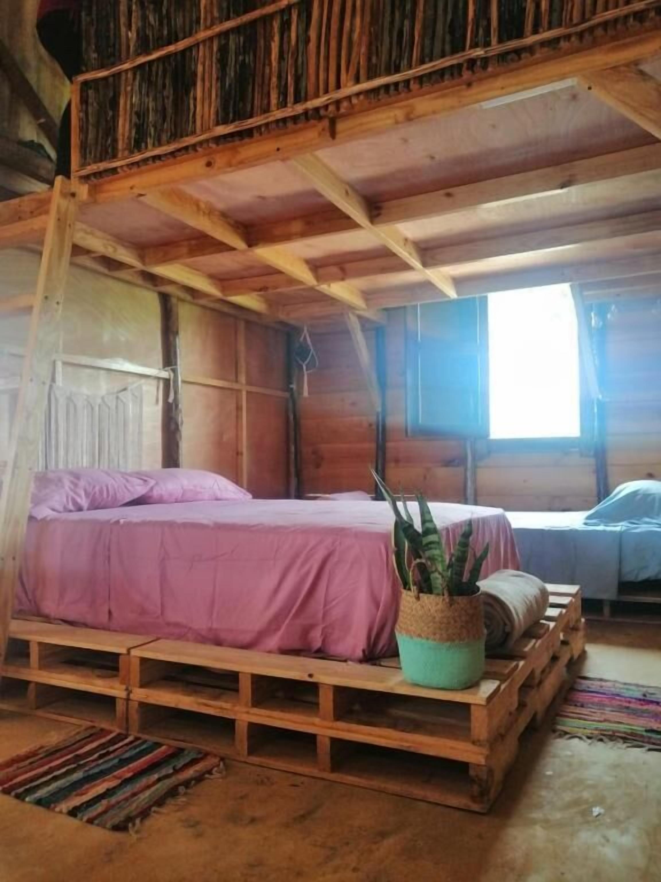 Una cabaña de madera rústica con un dormitorio en el altillo, una cama individual con colcha rosa y una pequeña mesa con una planta.