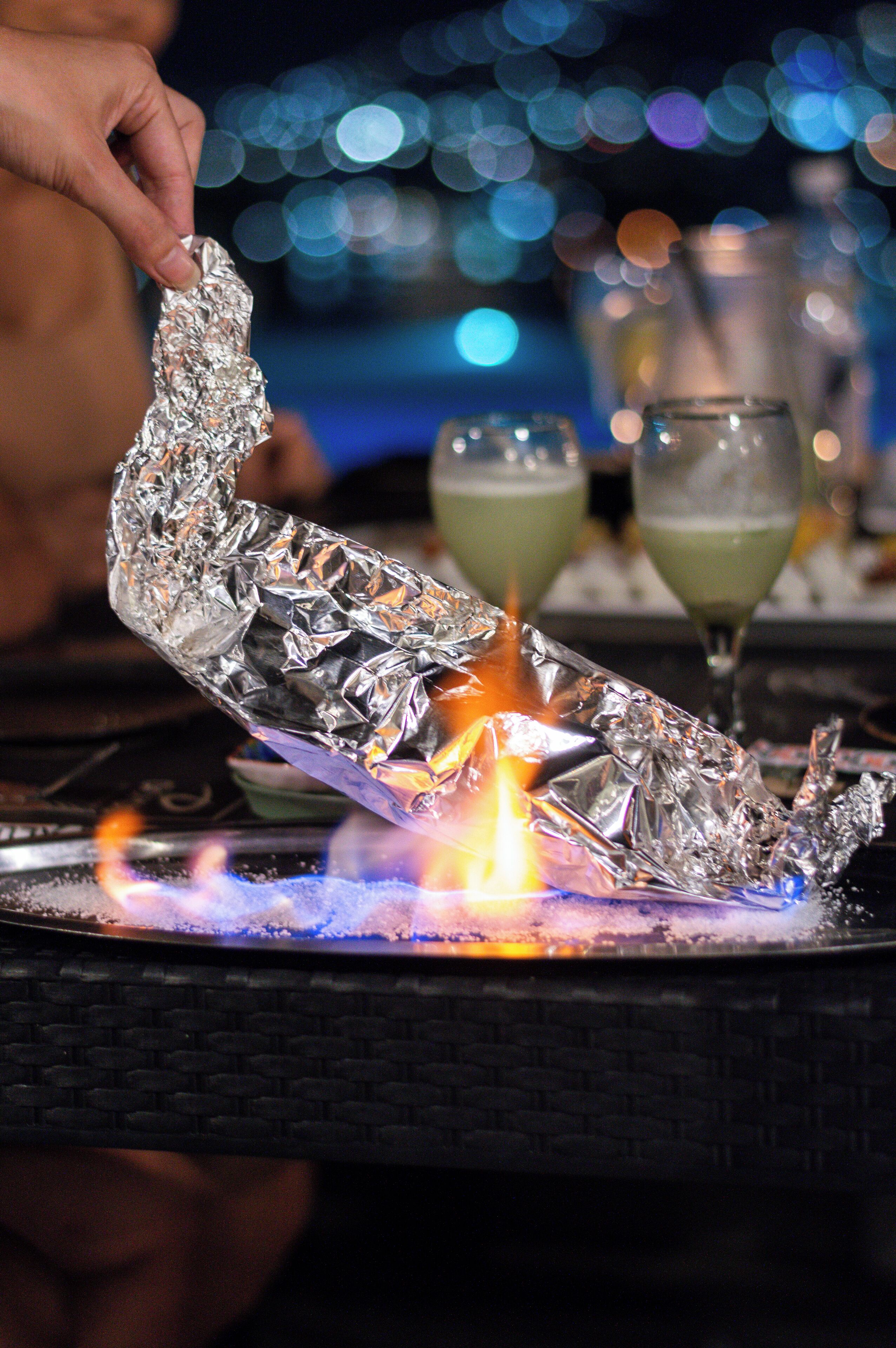 A hand holding a foil-wrapped object over a lit flame, with glasses and a blurred background.