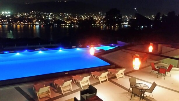 A nighttime view of a rooftop pool area with lounge chairs and tables, overlooking a cityscape with lights reflecting on the water. - Domus Lake Resort (Villa Carlos Paz)
