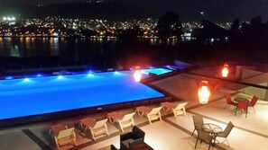 A nighttime view of a rooftop pool area with lounge chairs and tables, overlooking a cityscape with lights reflecting on the water. - Domus Lake Resort (Villa Carlos Paz)