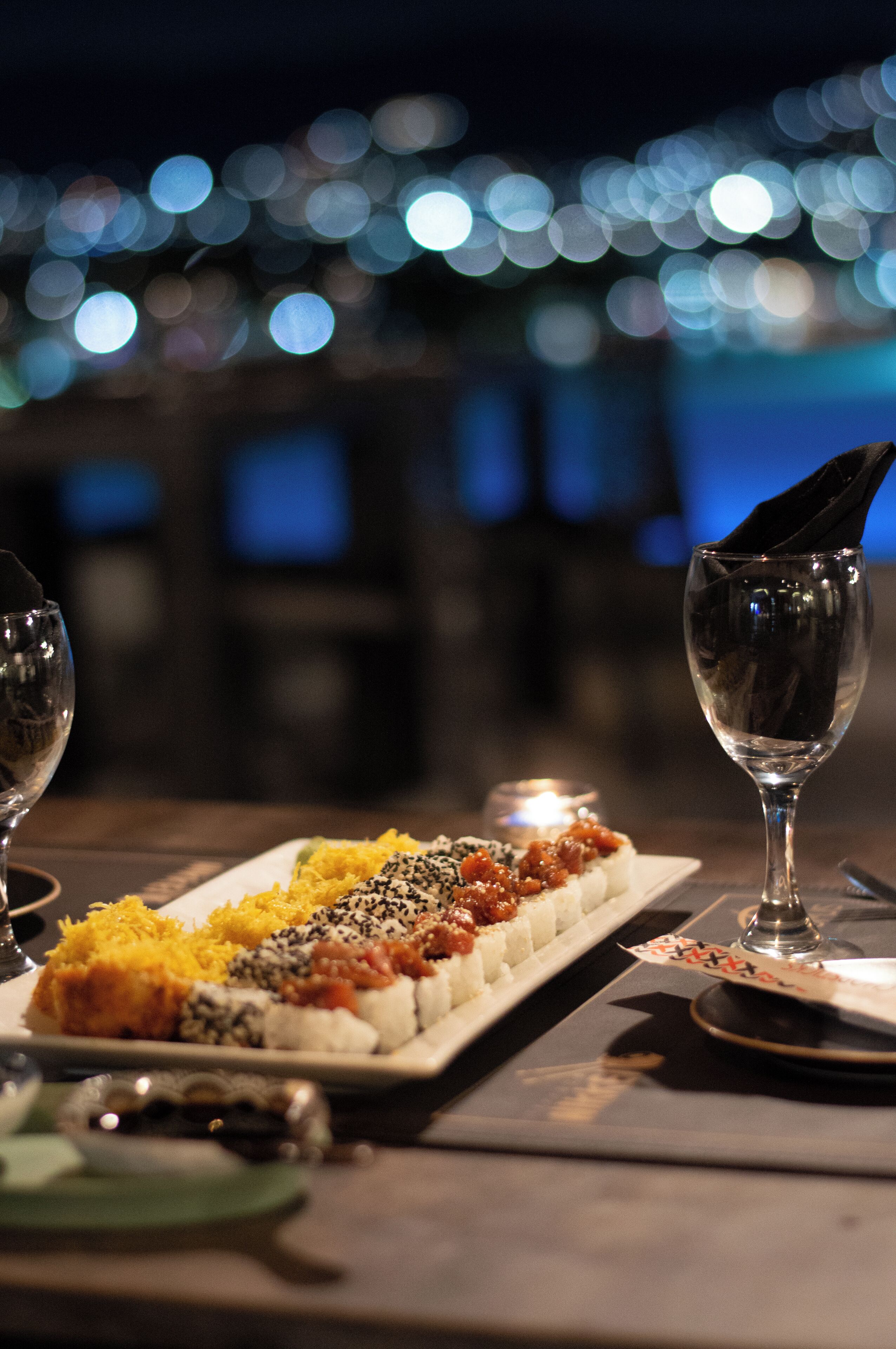 A dining table with a sushi platter, two wine glasses, and a candle.