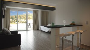 A modern hotel room with a large sliding glass door leading to a balcony, a sofa, a small kitchen area with bar stools, and a bed with a white duvet. - Domus Lake Resort (Villa Carlos Paz)