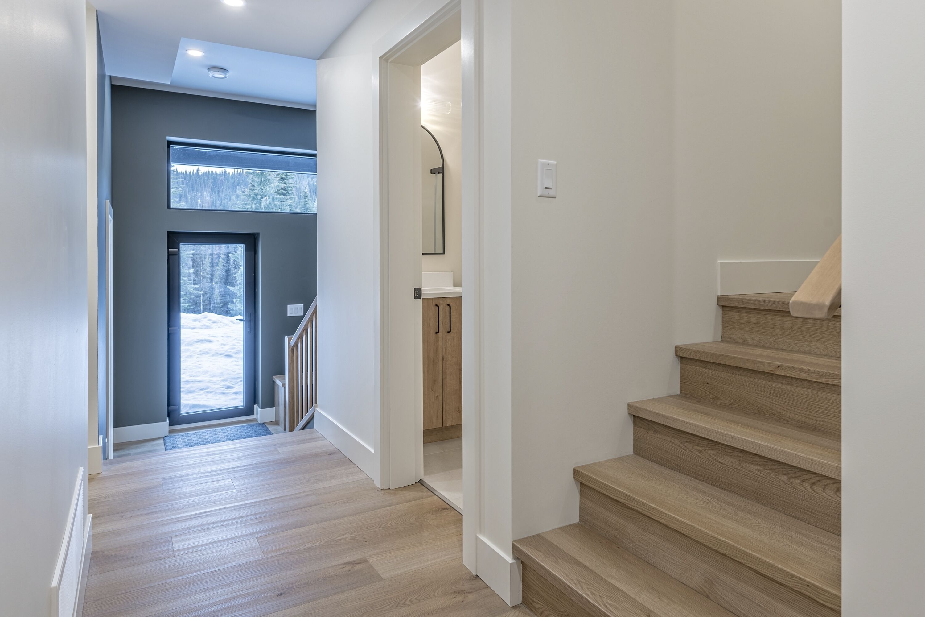 A modern interior with a wooden staircase, a door leading to a room, and a view of a snowy landscape through a large window.
