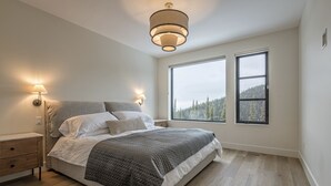 A bedroom with a large bed, two bedside lamps, a nightstand, and a window with a view of trees. - 5 bedroom home with direct ski access, sauna, private laundry and garage (Silver Star Mountain)