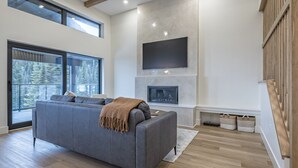 A modern living room with a grey sofa, a fireplace, and large windows offering a view of trees. - 5 bedroom home with direct ski access, sauna, private laundry and garage (Silver Star Mountain)