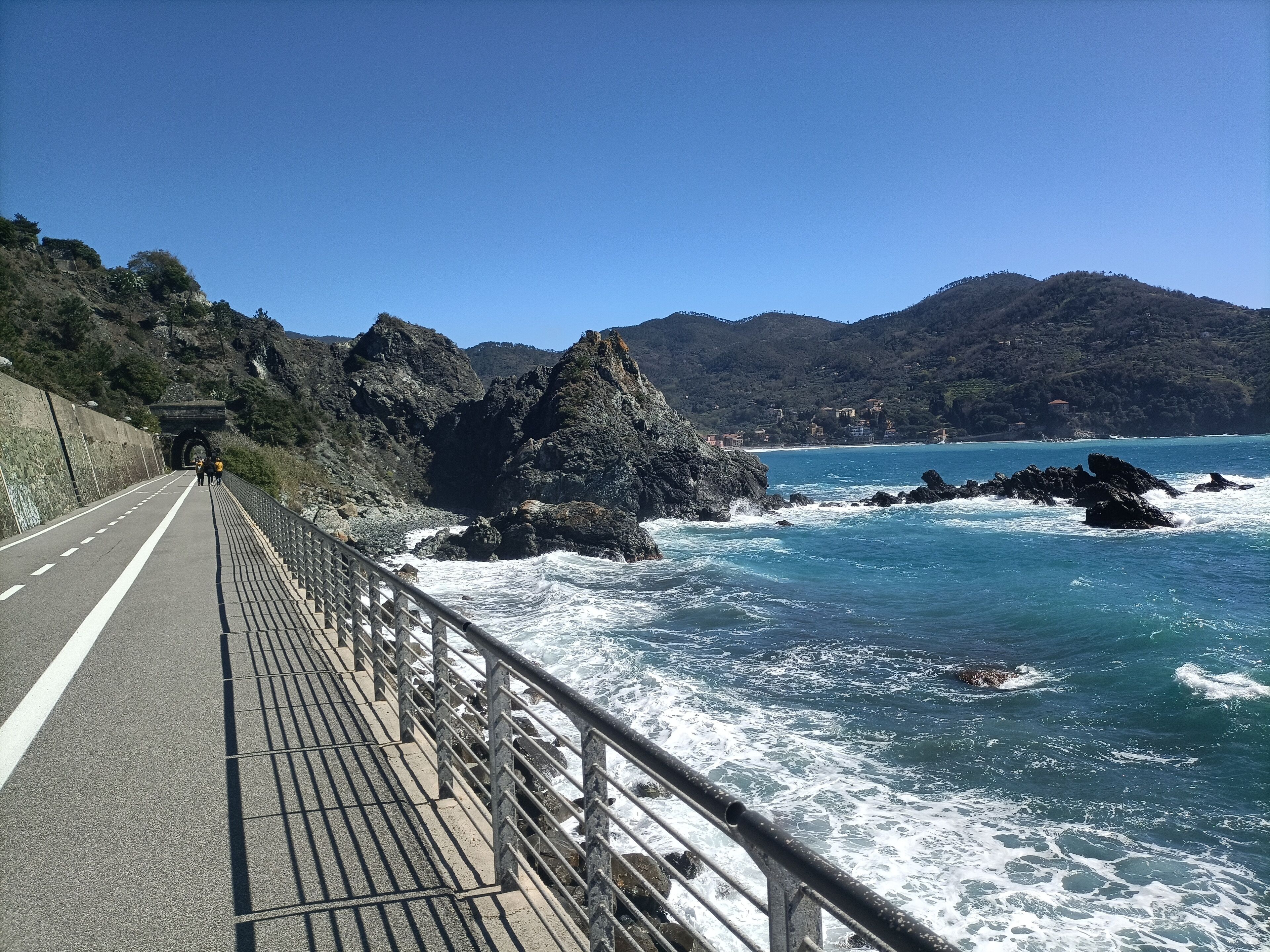 A coastal road with a metal guardrail, rocky cliffs, and waves crashing against rocks.