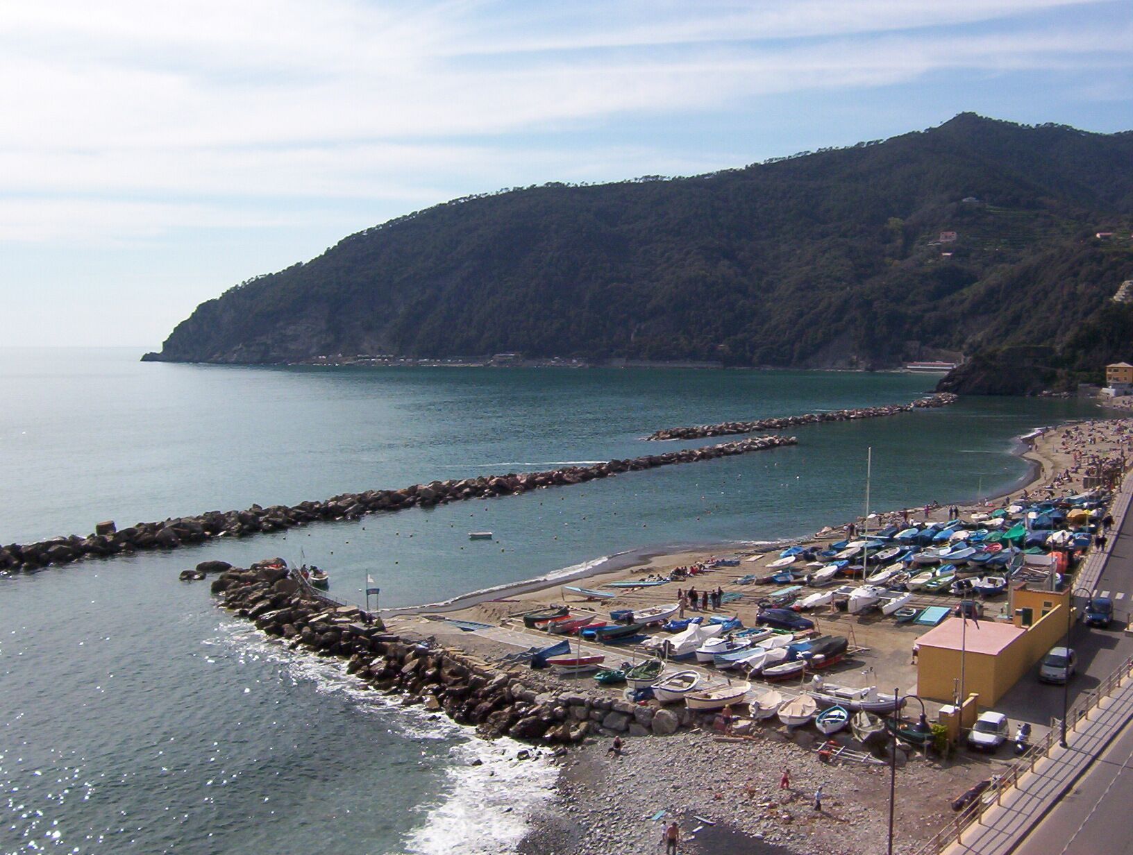 A coastal area with boats docked along the shore, a rocky breakwater, and a mountainous backdrop.