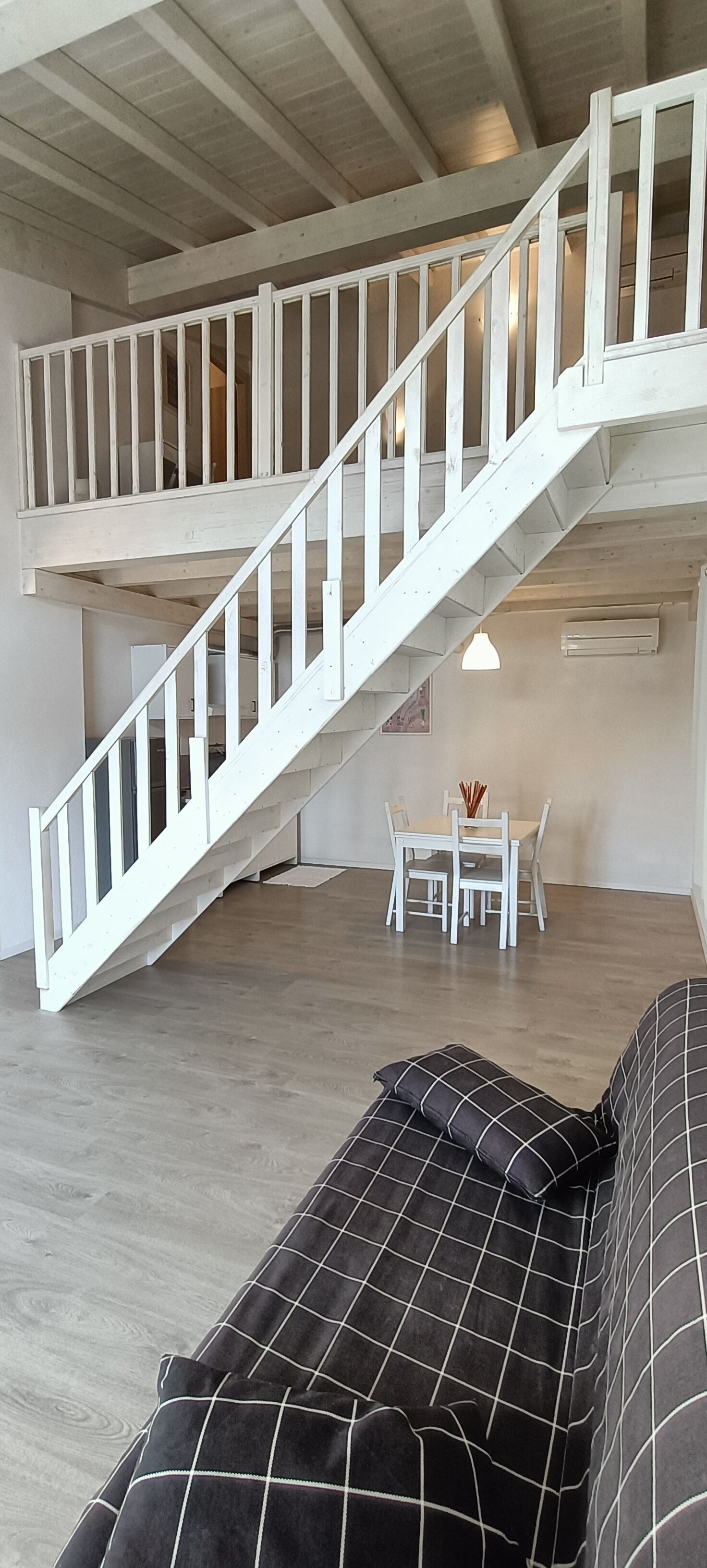 A spacious living area with a wooden staircase leading to an upper level, a dining table with chairs, and a black and white patterned sofa.