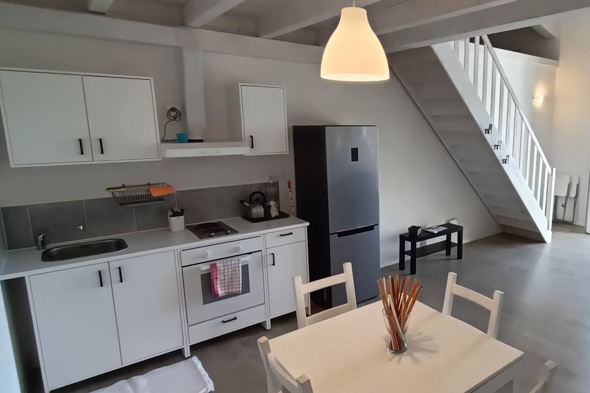 A modern kitchen with white cabinets, a black refrigerator, and a dining area with a table and chairs. A staircase leads to a balcony with a view of greenery.