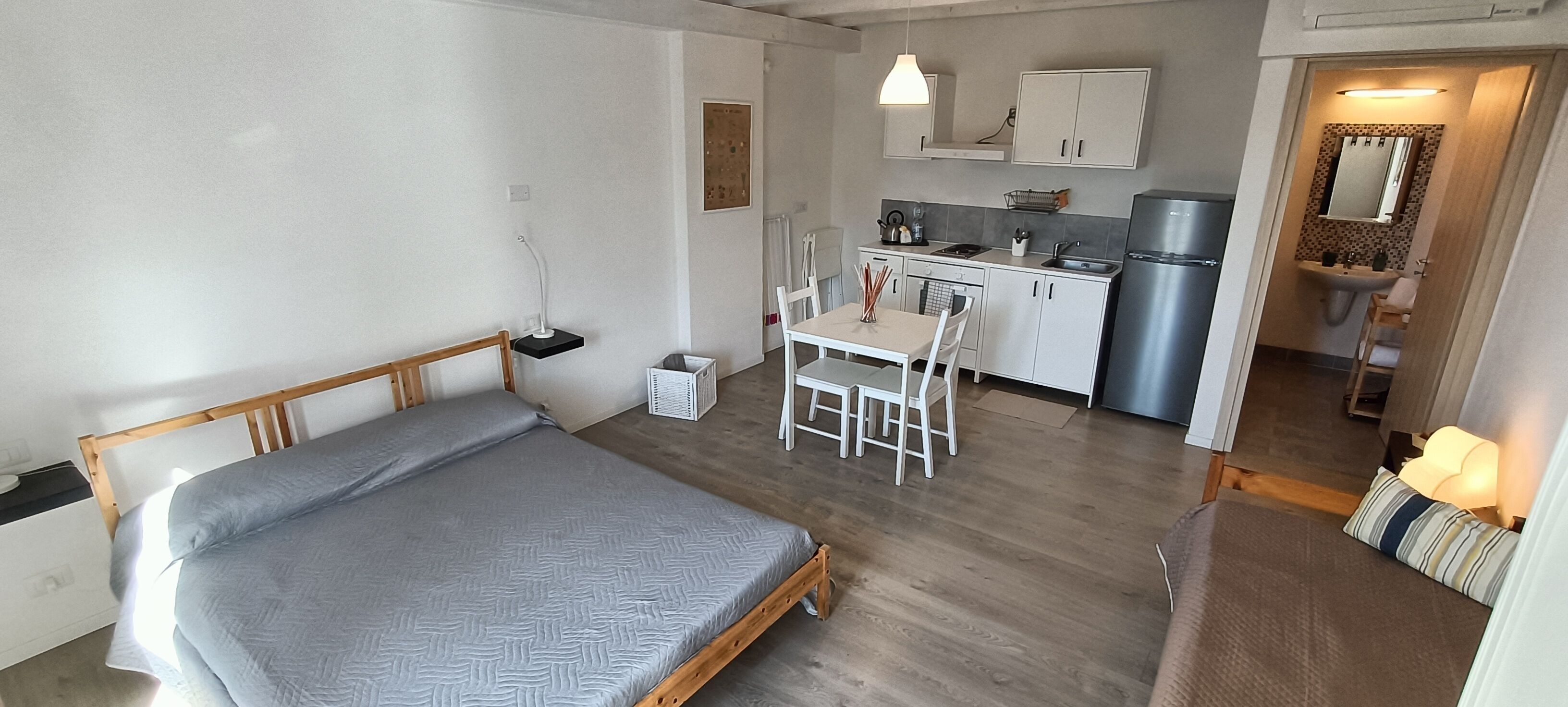 Ein kleines Schlafzimmer mit einem Einzelbett, einem kleinen Tisch und einem Stuhl. Angrenzend eine Küchenzeile mit Kühlschrank, Herd und Spüle.