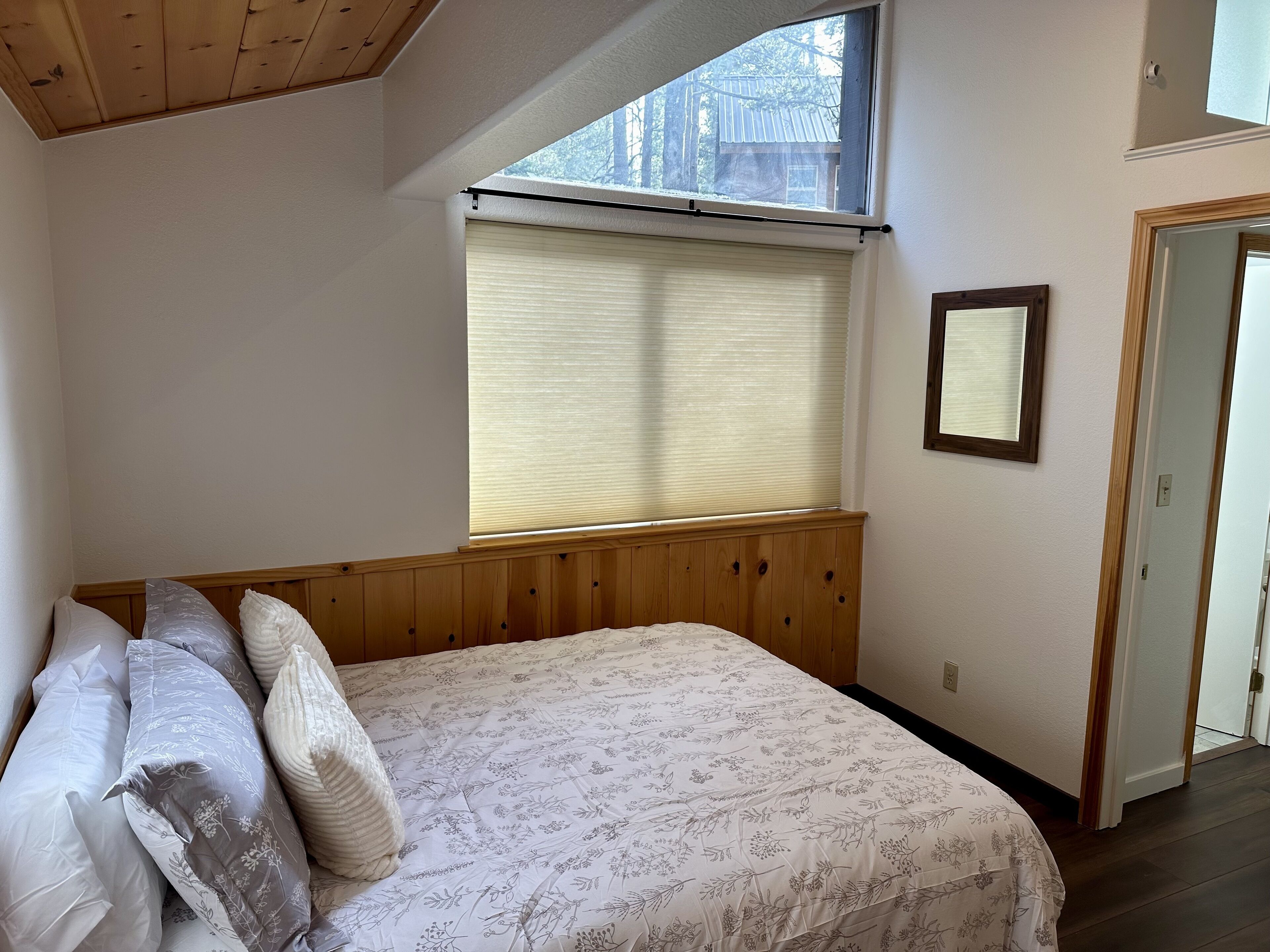 Un dormitorio con cabecera de madera, un ventanal con persianas, una cama con ropa de cama floral y una puerta que da a otra habitación.