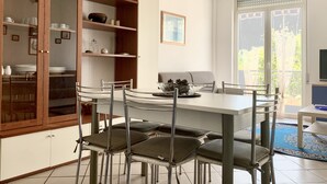 Een eetgedeelte met een witte tafel en stoelen, een houten kast met glazen deuren, en een groot raam met uitzicht op groen.