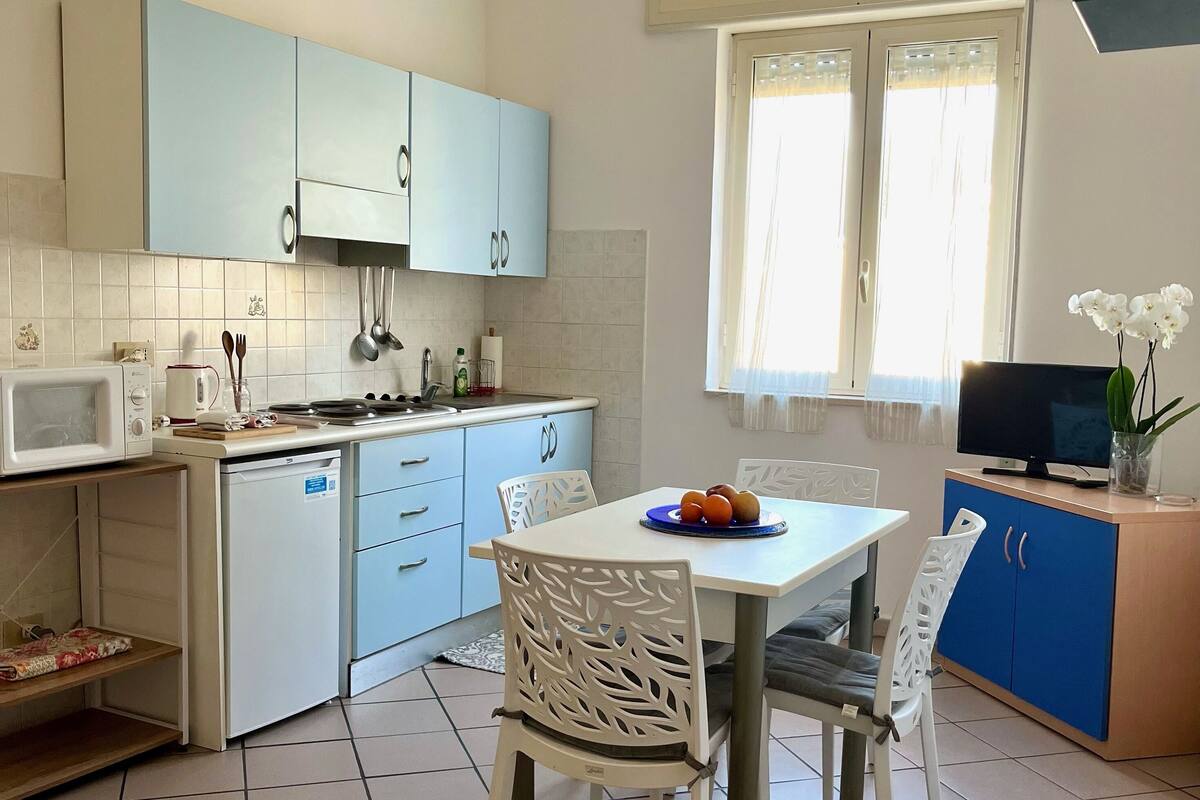 A compact kitchen with a dining area, featuring a microwave, stove, and sink. There is a dining table with chairs and a small TV on a cabinet.