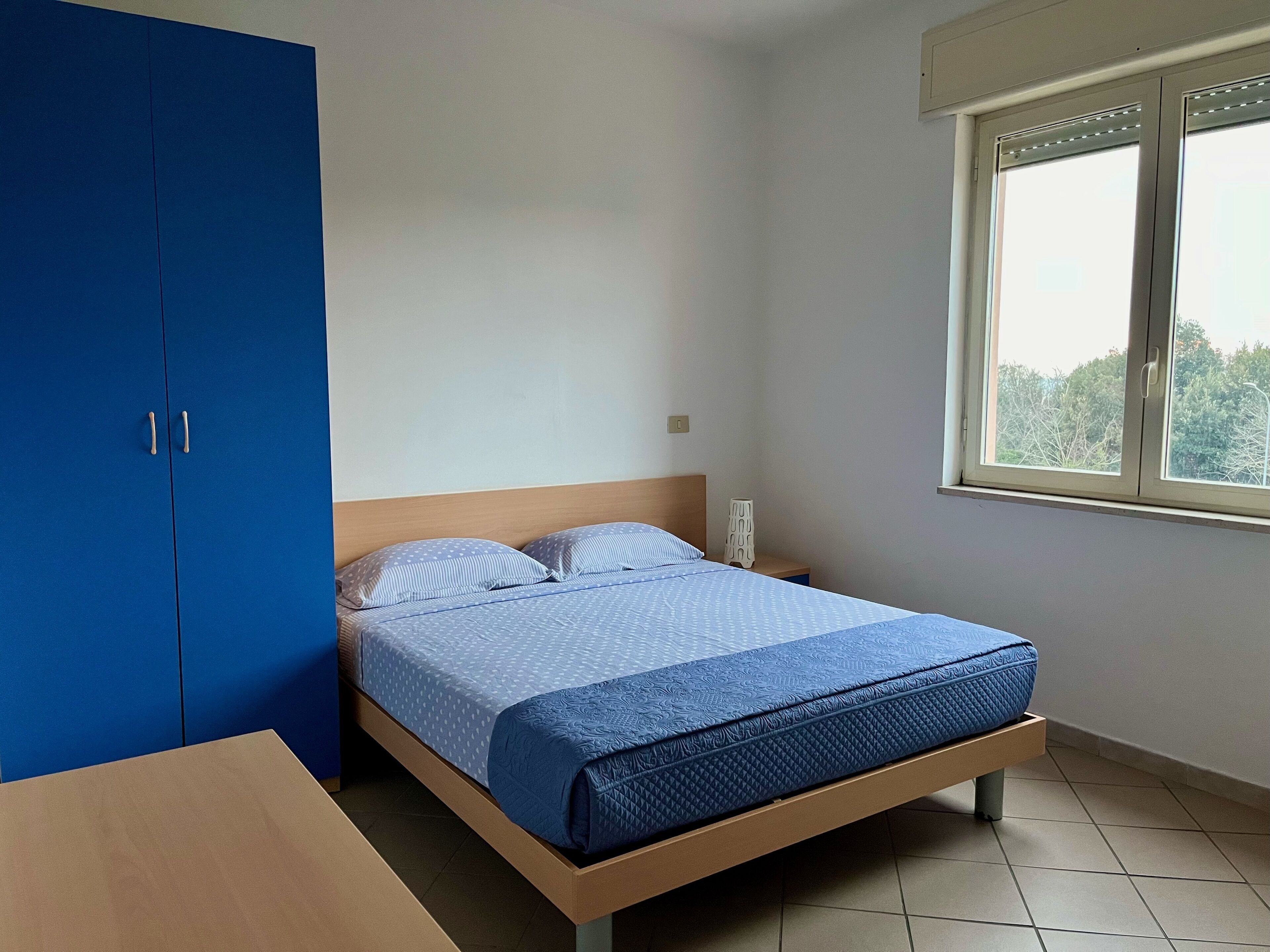 Una camera da letto con un letto, un comodino in legno, un armadio blu e una finestra vista alberi.