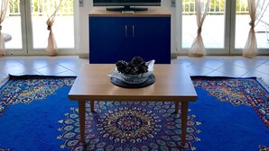 Een woonkamer met een flatscreen televisie op een blauwe kast, een houten tafel met een decoratieve schaal, en een blauw tapijt met patroon.