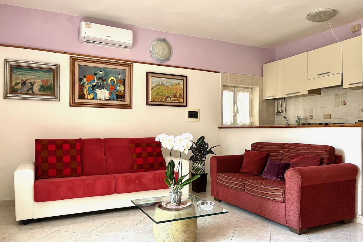 A living room with a red sofa, a glass coffee table, and a potted plant. There are framed pictures on the wall and a kitchen in the background.