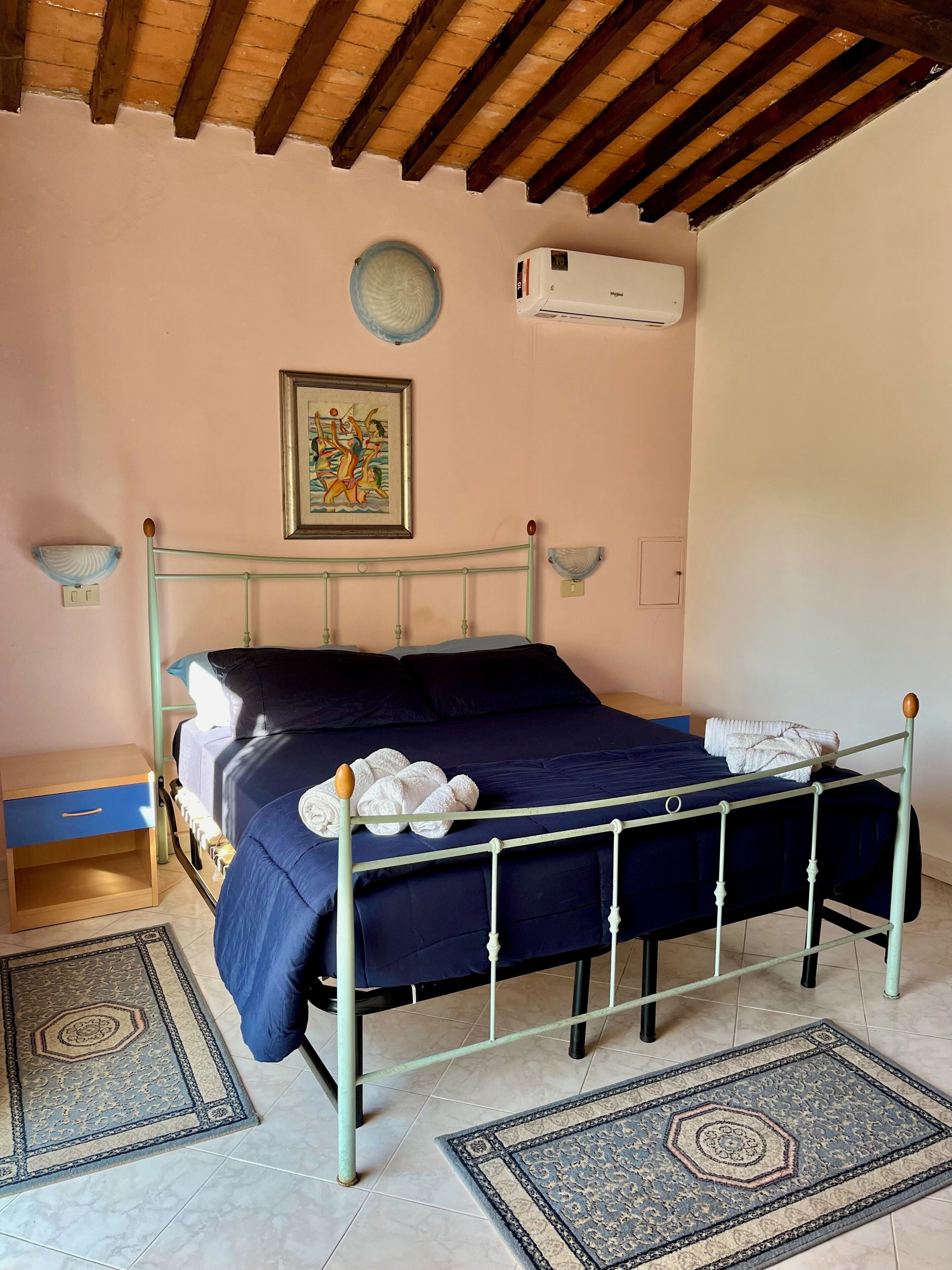 Une chambre à coucher comprenant un lit, une table de chevet, un climatiseur fixé au mur, un tableau encadré et un vase décoratif.