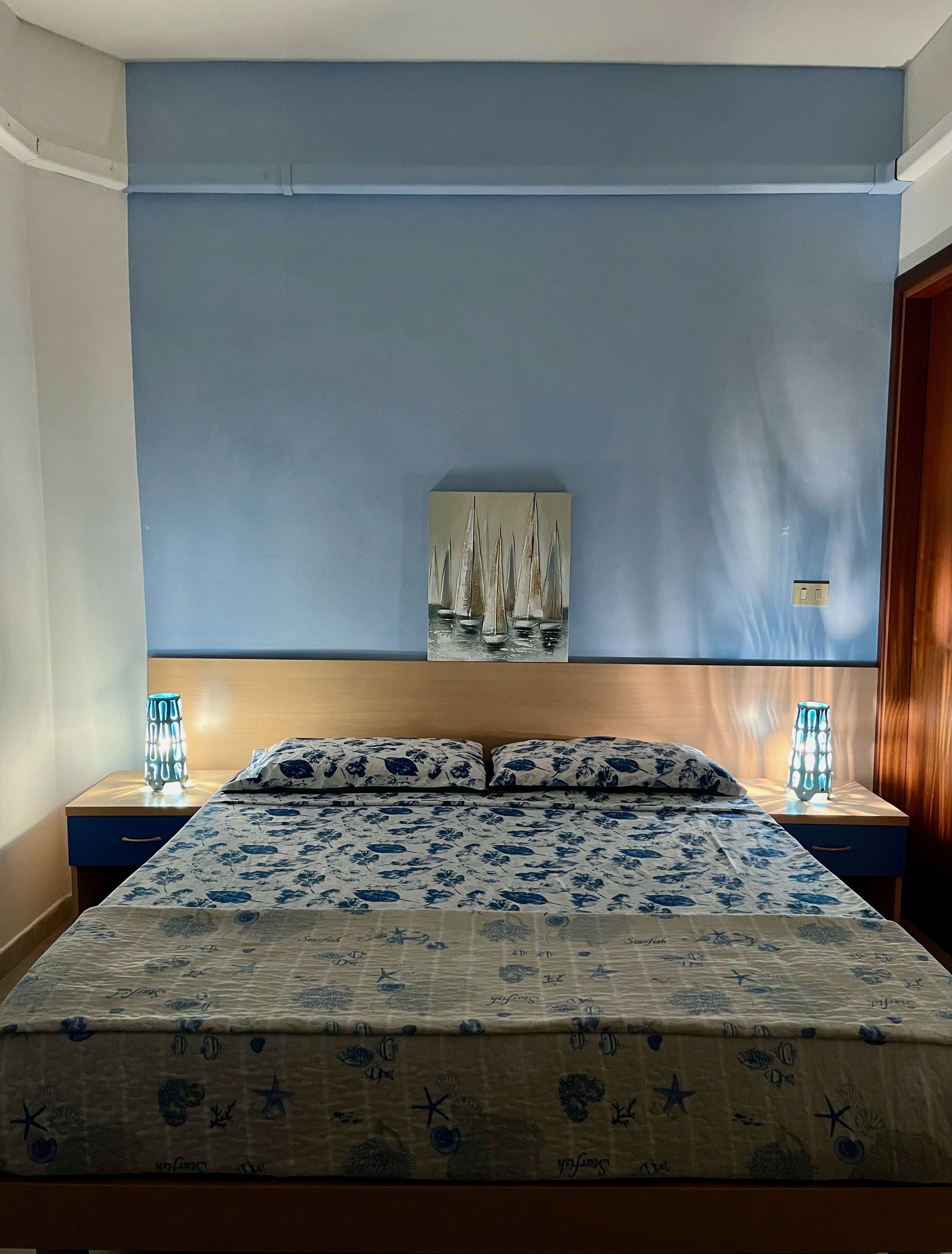 A bedroom with a bed, two bedside tables, a nightstand, a wall-mounted painting, and a night lamp.