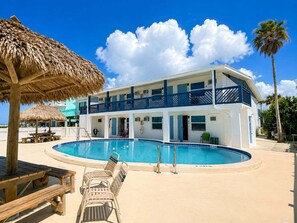 Um edifício de dois andares com piscina, guarda-sol de palha e uma cadeira de praia.