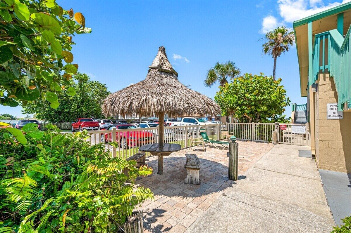 A thatched-roof structure with a table and chairs, surrounded by greenery and a parking area with cars.