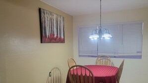 Comedor con una mesa dispuesta para cuatro, sillas de madera, mantel rojo, un cuadro en la pared y una ventana con persianas.