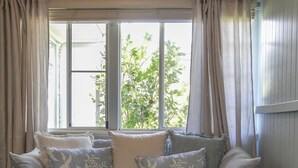 Ein gemütliches Zimmer mit einem grauen Sofa, verziert mit floralen Kissen, einem kleinen runden Tisch und einem Fenster mit beigen Vorhängen.